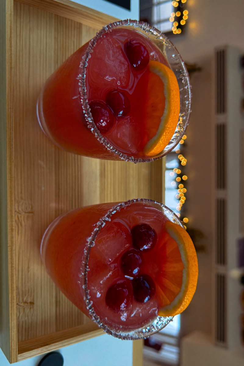 Two cranberry orange margaritas on a wooden tray with Christmas lights behind